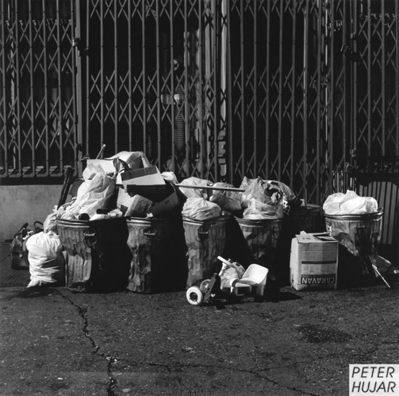 Fileira de latas de lixo cheias em frente a portões de ferro fechados.