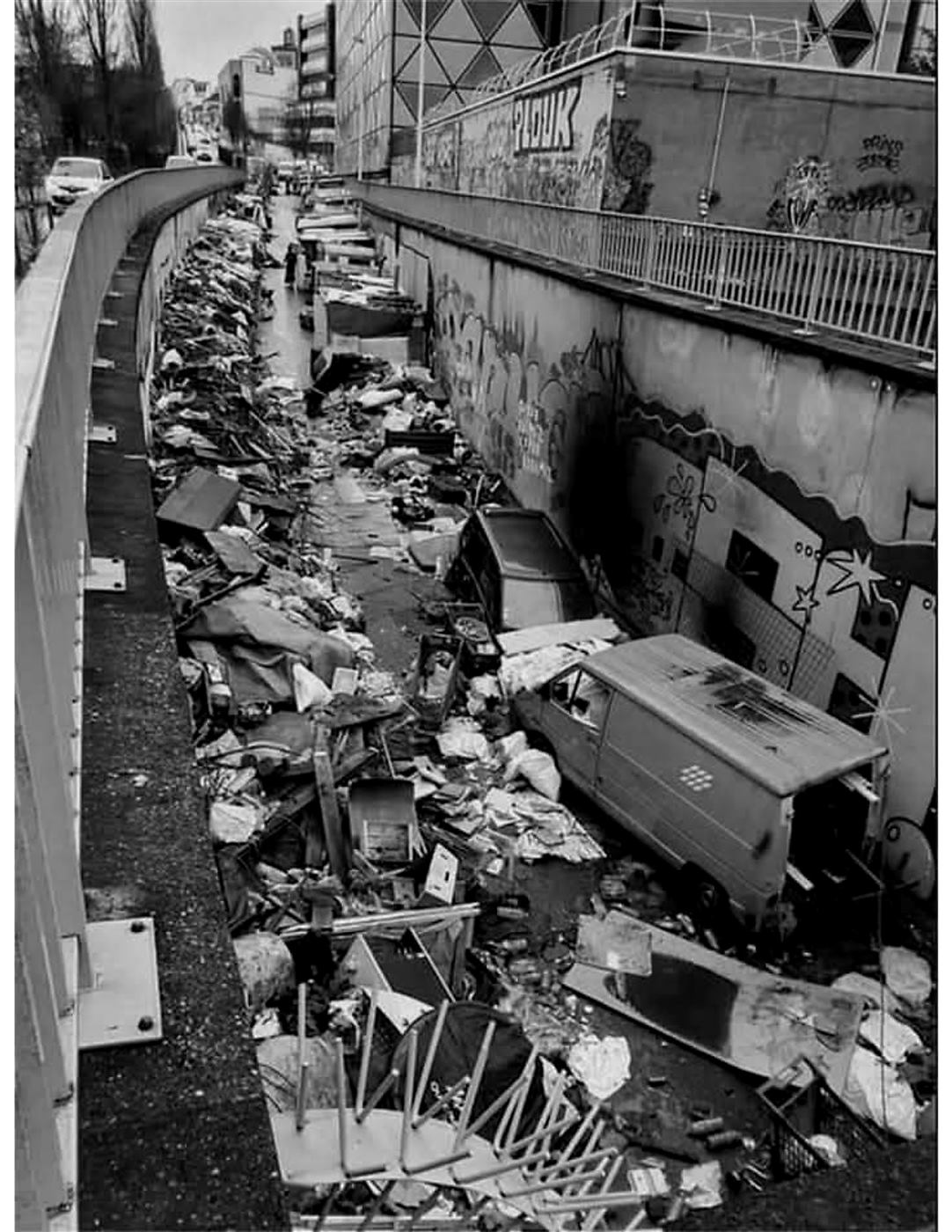 Rua estreita tomada por entulho, móveis quebrados e carros abandonados.